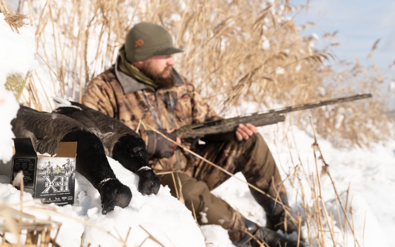 hunter sitting in the snow behind dead geese and HEVI-XII packaging